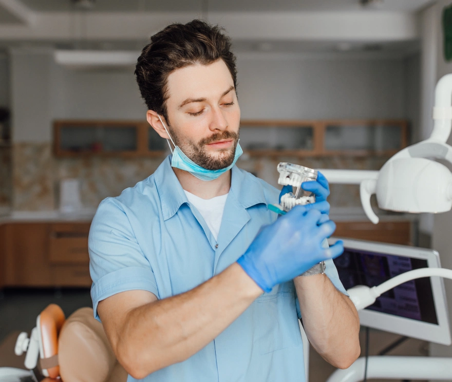 Male dentist in blue uniform and gloves holding a dental model in a clinic.